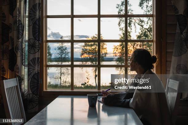 finland, lapland, young woman sitting at the window looking at a lake - abendstimmung natur ruhe stock-fotos und bilder