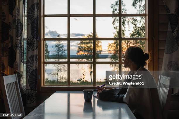finland, lapland, young woman sitting at the window looking at a lake - author stock pictures, royalty-free photos & images