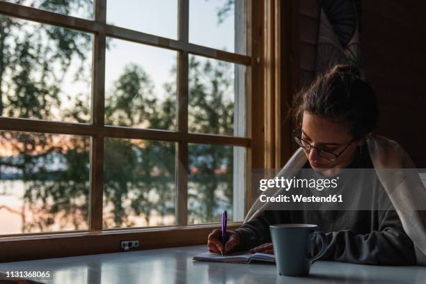 finland, lapland, young woman sitting at the window at a lake writing into diary - diary stock-fotos und bilder