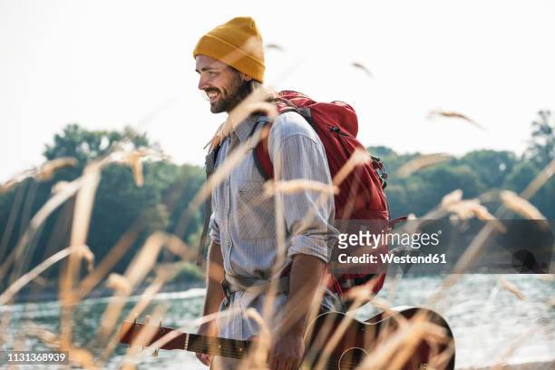 smiling young man with backpack and guitar at the riverside - naturbursche stock-fotos und bilder