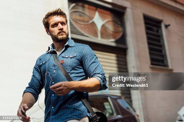 portrait of businessman using smartphone and earphones outdoors - crossbody bag stock pictures, royalty-free photos & images
