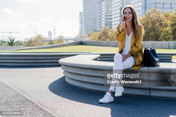 happy woman with bag and cell phone sitting on a bench in the city - business park stockfoto's en -beelden