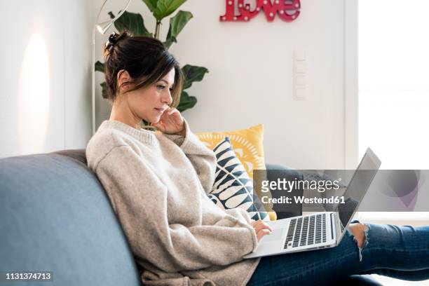 woman sitting on her couch, surfing the net, using laptop - download photos et images de collection