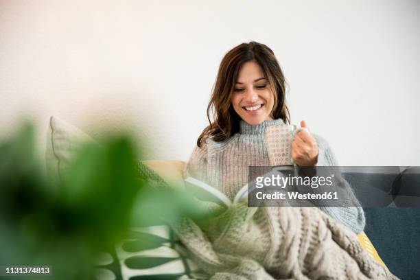 woman sitting on couch, wrapped in a blanket, reading book, drinking tea - hygge bildbanksfoton och bilder