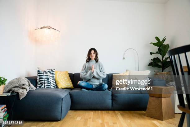 mature woman sitting cross-legged on couch with eyes closed, meditating at home - yoga kissen stock-fotos und bilder