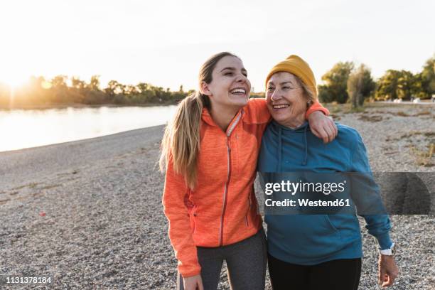 fit grandmother and granddaughter walking at the river with arms around, having fun - granddaughter stockfoto's en -beelden