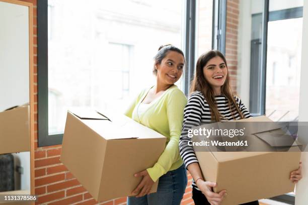 portrait of two smiling young women carrying cardboard boxes in new apartment at the window - student flat stock pictures, royalty-free photos & images