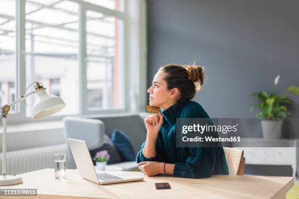 profile of pensive woman with laptop and credit card sitting at desk at home looking out of window - casual woman pensive side view stockfoto's en -beelden