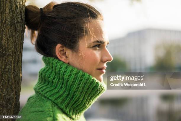 profile of relaxed woman wearing green turtleneck pullover leaning against tree trunk - mock turtleneck stock-fotos und bilder