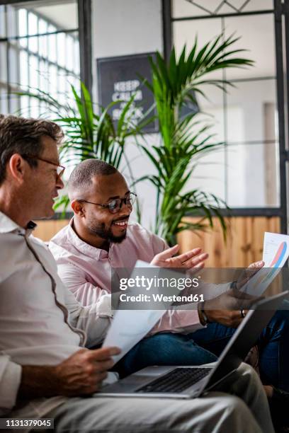 two businessmen using laptop and discussing documents on sofa in loft office - business people and paper imagens e fotografias de stock