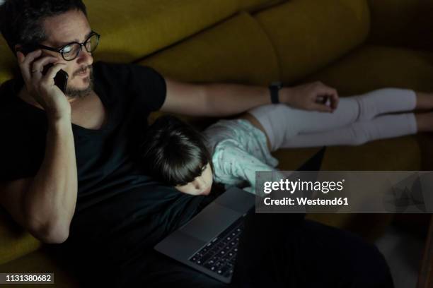 father talking on cell phone on couch at night with daughter lying next to him - daughters of darkness - fotografias e filmes do acervo