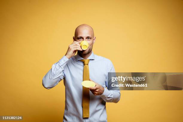 portrait of bald businessman drinking cup of coffee against yellow background - bald man foto e immagini stock