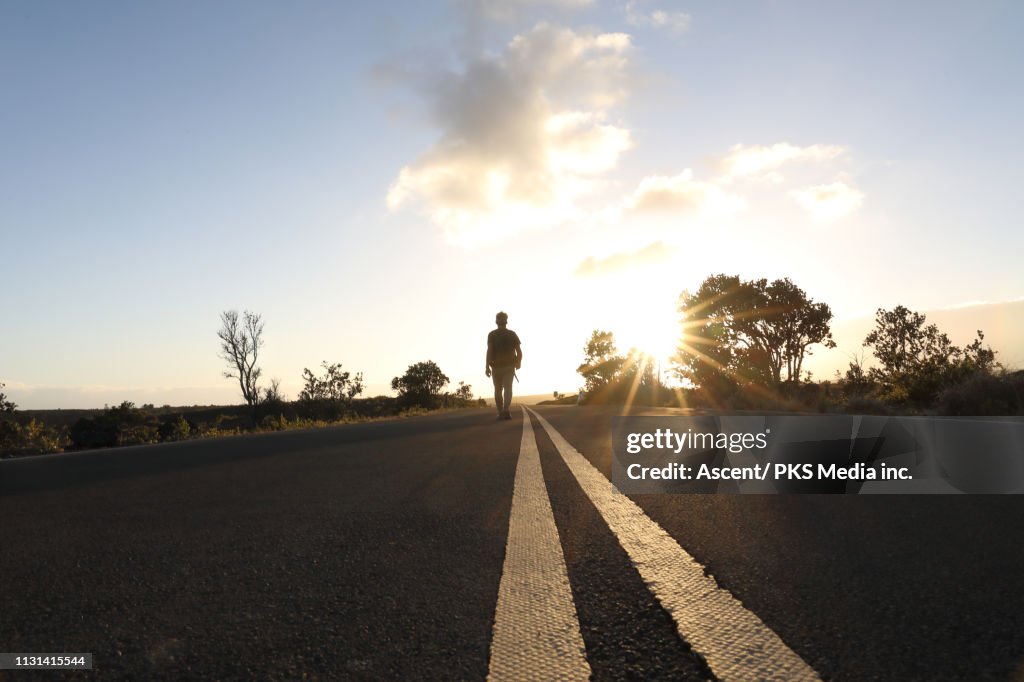 Man walks down centre line of highway into the sunset