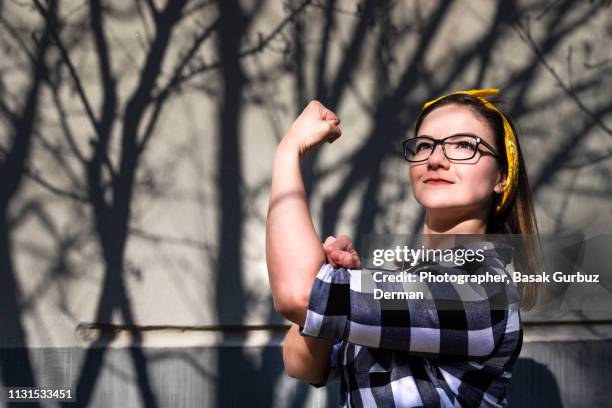 portrait of a young, powerful, beautiful woman wearing glasses and a bandana, flexing her bicep muscle - womens day stock-fotos und bilder