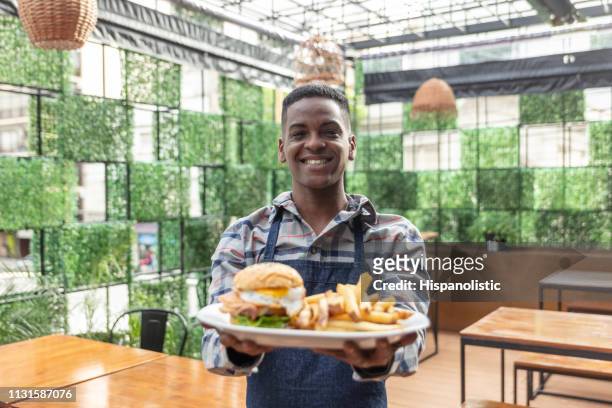 serveur mâle d'afro-américain dans un restaurant retenant une assiette avec le burger et les frites tout en faisant face souriant de caméra - burger portrait photos et images de collection