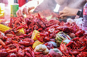 Boiled crawfish and vegetables piled on red checked tablecloth with eating tray and arm of person eating bokeh behind - shallow focus