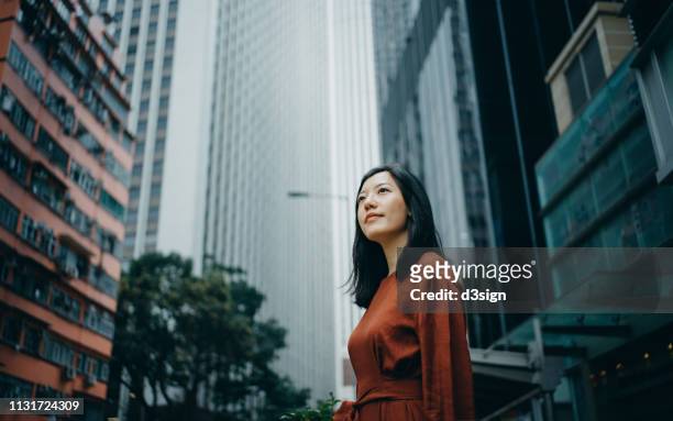 low angle portrait of confidence young woman standing against highrise city buildings in city - förväntan bildbanksfoton och bilder