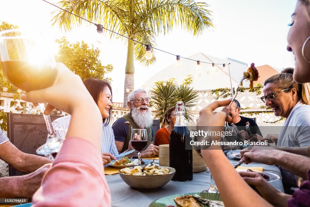 Happy family doing a dinner during sunset time outdoor - Group of diverse friends having fun dining together outside - Concept of lifestyle people, food and weekend activities