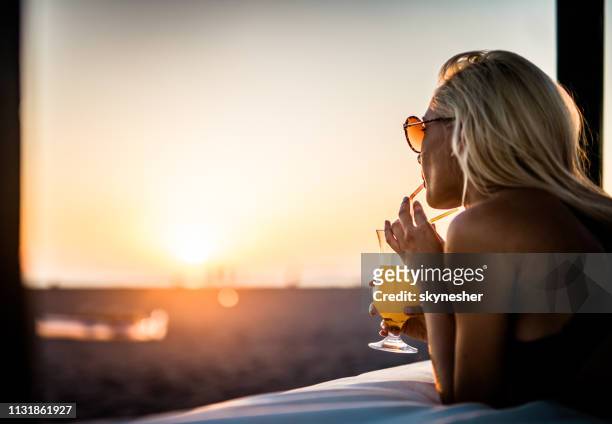 junge frau trinkt bei sonnenuntergang sommercocktail auf dem strandbett - orange juice stock-fotos und bilder