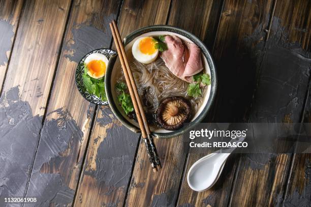 Traditional Japanese Noodle Soup with shiitake mushroom. Egg. Sliced beef and greens served in ceramic bowl with wooden chopsticks and white spoon....