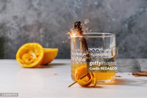 Glass of Scotch Whiskey orange juice alcohol cocktail with swirled orange peel and smoking cinnamon sticks standing on white marble table with golden...