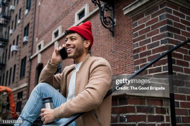 young man sitting on stoop talking on mobile phone - red hat stock pictures, royalty-free photos & images