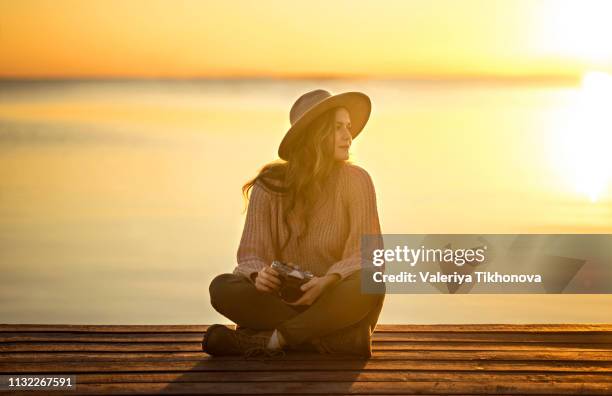young woman with camera sitting on pier at sunset - ponton photos et images de collection