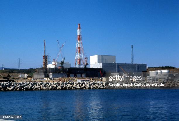 Wide shot of the Fukushima Daiichi Nuclear Power Plant, under construction on a sunny day, with a seawall and the Pacific Ocean in the foreground,...