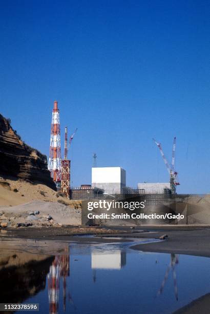 Fukushima Daiichi Nuclear Power Plant, under construction on a sunny day, reflected in a pool of water cutting through sand in the foreground,...