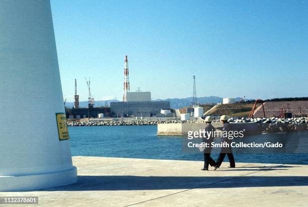 Two men wearing construction helmets walk on a concrete pier, on a sunny day, with the Fukushima Daiichi Nuclear Power Plant in the background,...