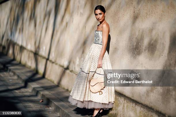 Geraldine Boublil wears a Dior pleated white off-shoulder dress with printed features, polka dots, a Dior Saddle bag, outside Dior, during Paris...