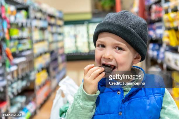 glimlachend drie jaar oud jongetje zit in een winkelwagentje bij een supermarkt gangpad met een chocolade cookie - years of the conran shop stockfoto's en -beelden