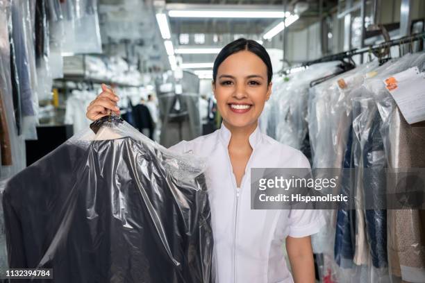 beautiful employee at a laundromat holding a dry cleaned outfit covered with a bag while facing camera smiling - lavandaria imagens e fotografias de stock