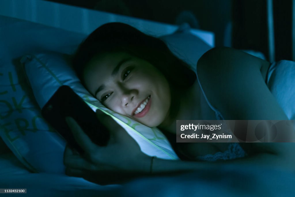 Mulher sorridente usando o telefone móvel enquanto deitada na cama à noite