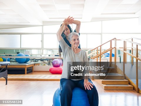 Senior woman sitting on exercise ball with physical therapist behind Senior woman sitting on exercise ball with physical therapist behind
