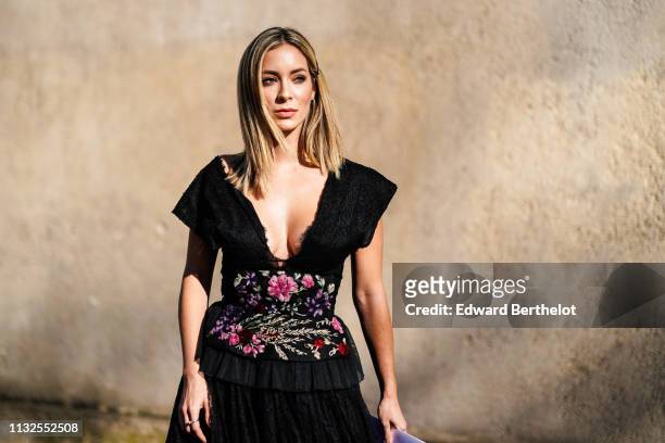 Guest wears a hair clip, earrings, a low-cut black lace ruffled dress with colorful floral embroideries on the waist, outside Dior, during Paris...
