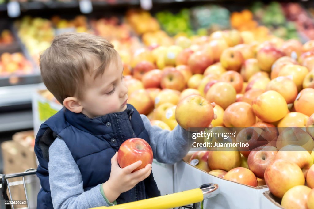 Three years old little child sitting in a shopping cart buying apples at a supermarket aisle
