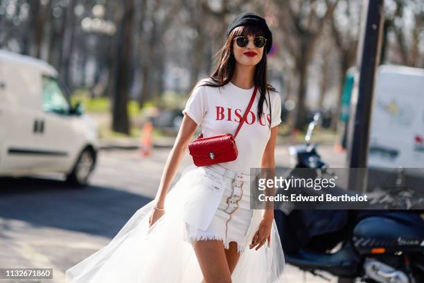 Gabrielle Caunesil wears a beret, sunglasses, a white t-shirt with red printed word "BISOUS", a red leather shoulder strapped Bulgari bag, white...