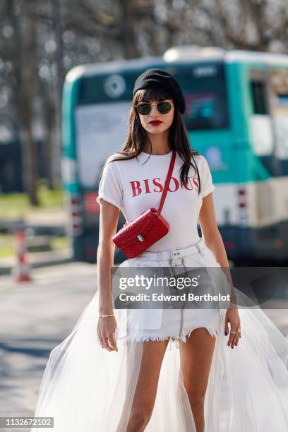 Gabrielle Caunesil wears a beret, sunglasses, a white t-shirt with red printed word "BISOUS", a red leather shoulder strapped Bulgari bag, white...