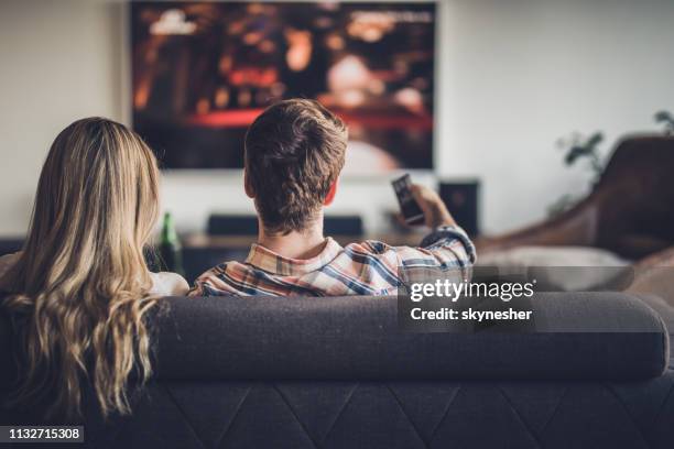 vue arrière d'un couple regardant la télévision tout en se relaxant sur le canapé à la maison. - regarder tv photos et images de collection