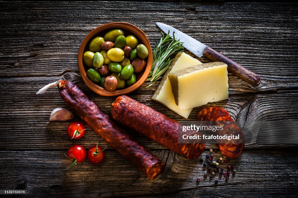 Comida española: queso manchego, chorizo español y aceitunas rodados desde arriba sobre mesa rústica de madera