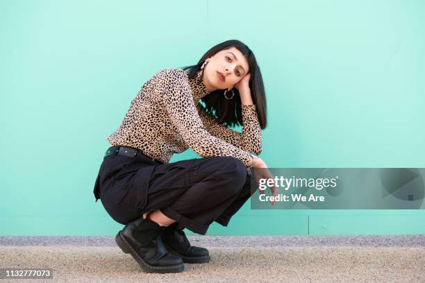 young woman crouching down in front of blue wall. - street style foto e immagini stock