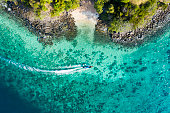 View from above, aerial view of a traditional long-tail boat sailing near a stunning barrier reef with a beautiful small beach bathed by a transparent and turquoise sea. Phi Phi Island, Thailand.