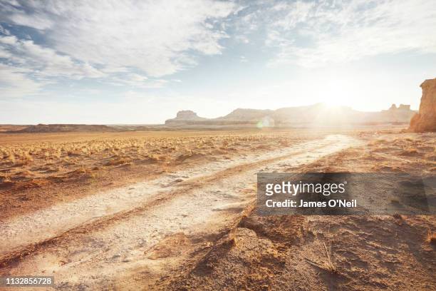 dirt road in arid desert landscape with distant cliffs and sunlight - terreno accidentato foto e immagini stock