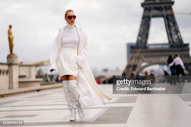 Elina Halimi wears sunglasses, a white coat, a white turtleneck top, a skirt, white leather thigh high boots, outside Ann Demeulemeester, during...