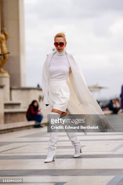 Elina Halimi wears sunglasses, a white coat, a white turtleneck top, a skirt, white leather thigh high boots, outside Ann Demeulemeester, during...