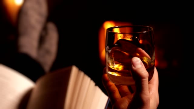 Man Reading Book And Drinking Glass Of Whisky Warming Feet By Flames Of Wood Burning Stove