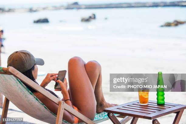 a young woman lying on a deck chair while touching a smartphone at the beach - sun deck stock pictures, royalty-free photos & images