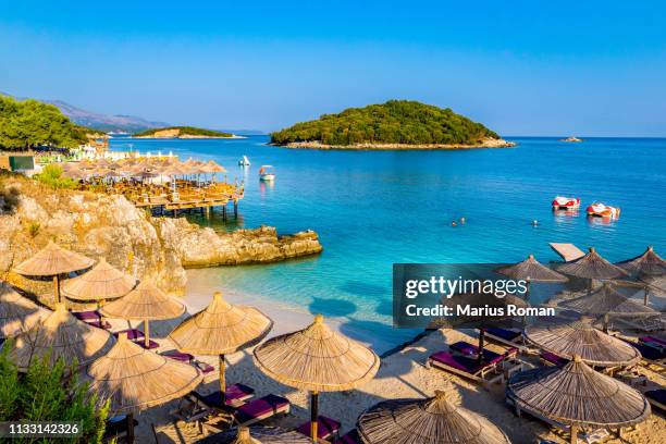 sunshade umbrellas, deckchairs and boats on the beautiful ksamil beach, vlore, ionian sea, albania, balkans, europe. - albanië stockfoto's en -beelden