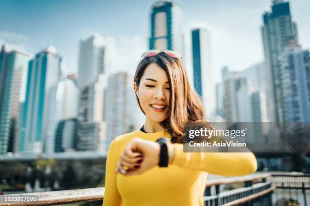 beautiful young sports woman checking smartwatch while exercising outdoors against urban cityscape - on time stock pictures, royalty-free photos & images